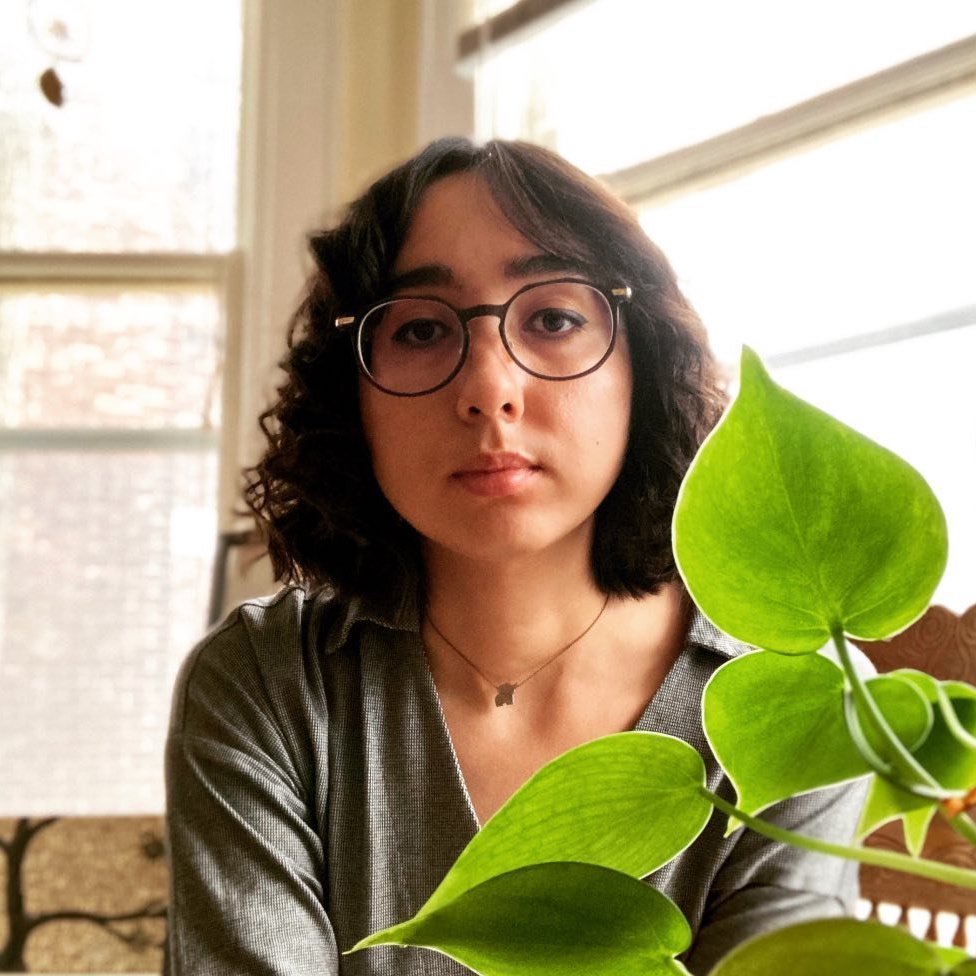 Serena smiling and sitting by a window with a plant in front of her. 