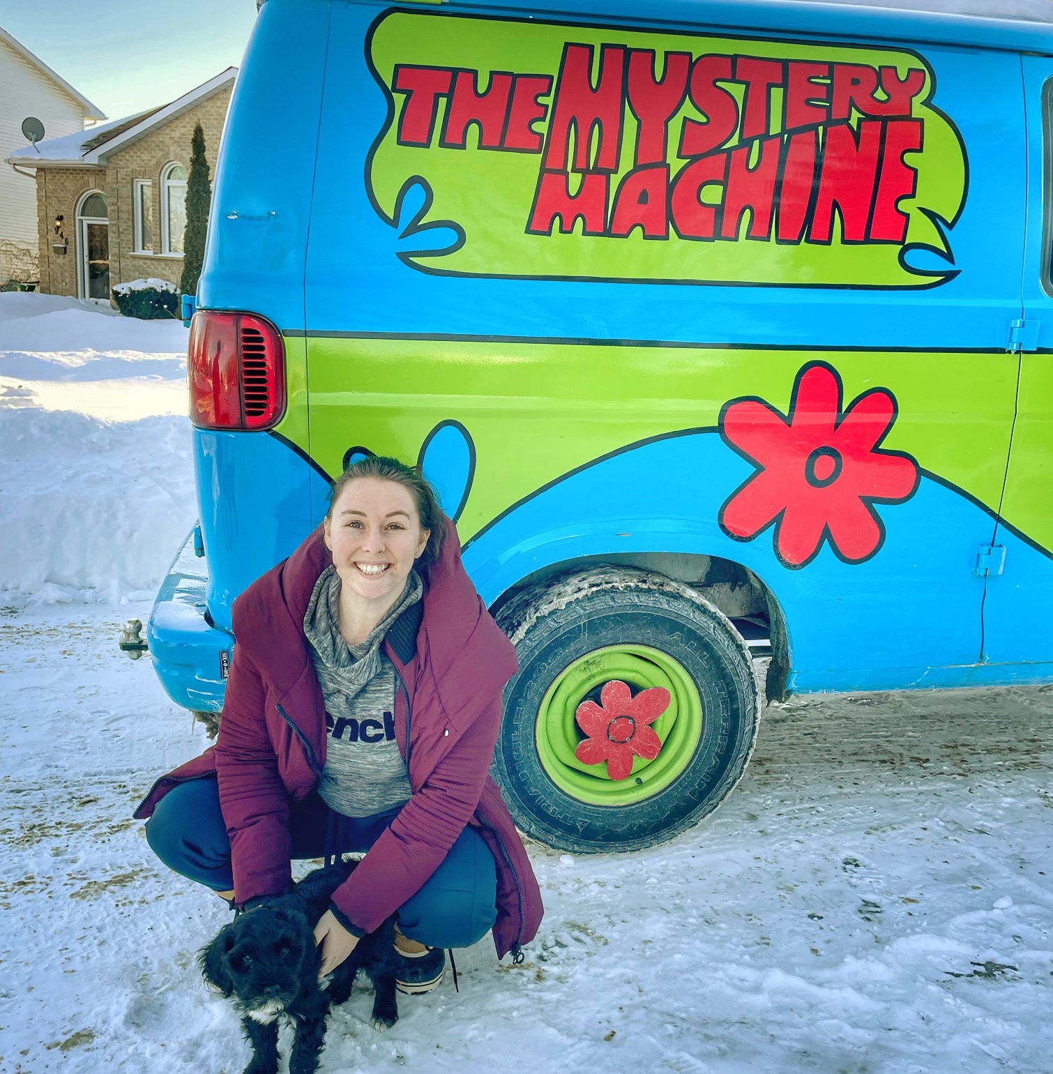 Casey kneeling with a small dog by the a van with 'The Mystery Machine' printed on it.