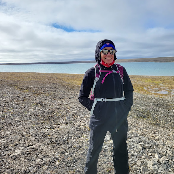 Brianna Lane smiling and standing in the Arctic, on a rocky beach in front of a large body of water.