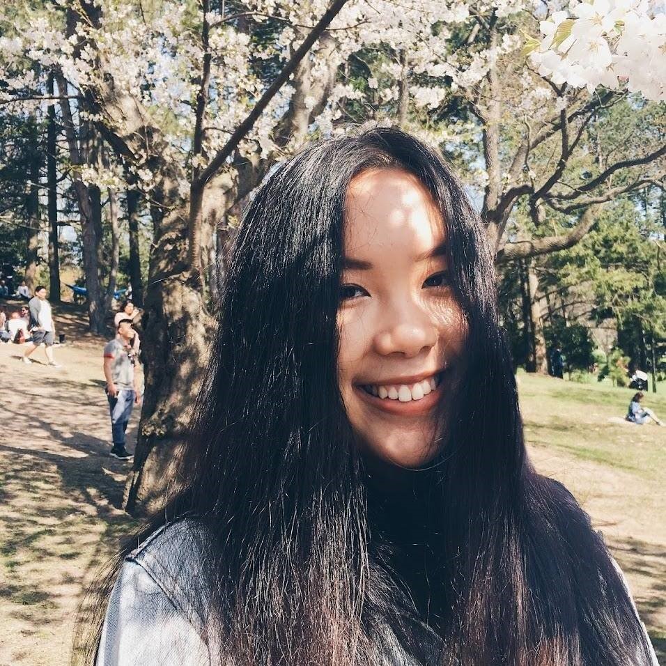 Cathy smiling in sunny park, in front of trees and grass. 