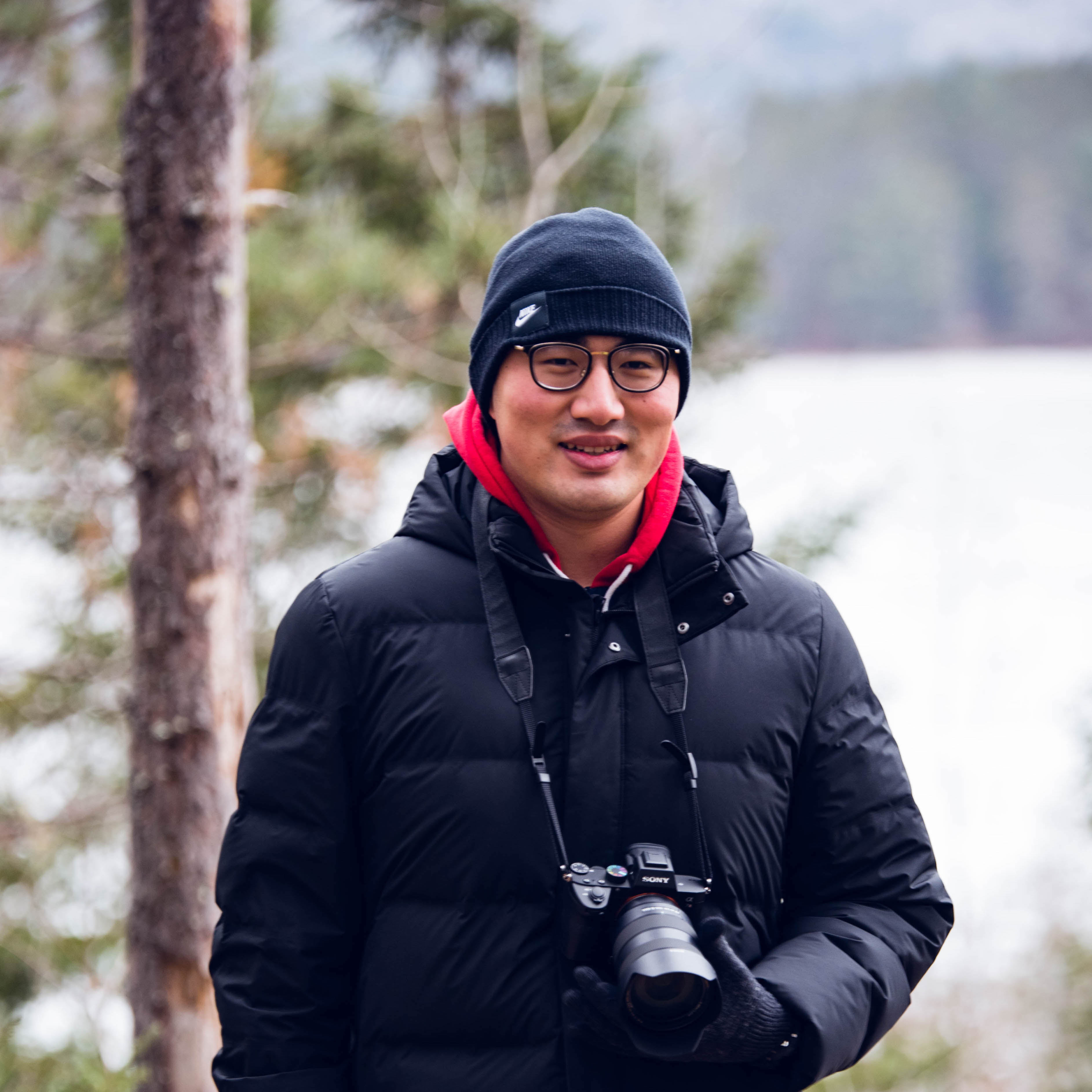 Jiye smiling and standing with a camera in front of some trees. 