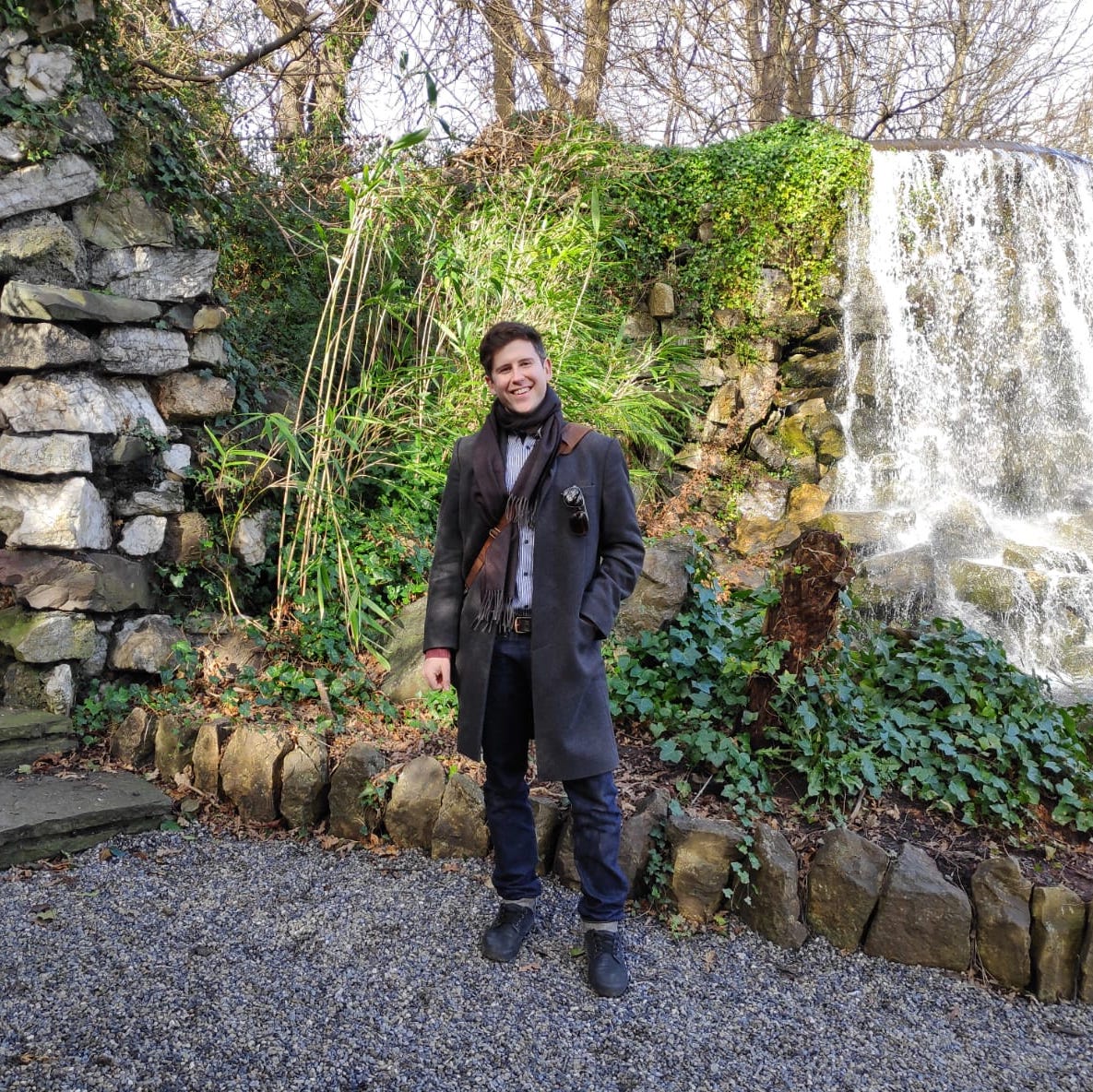 Michael smiling and standing on a stone path, in front of a small archway and waterfall. 