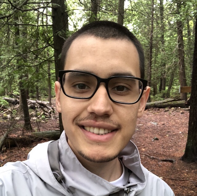 Alex smiling in grey jacket in a forest. 