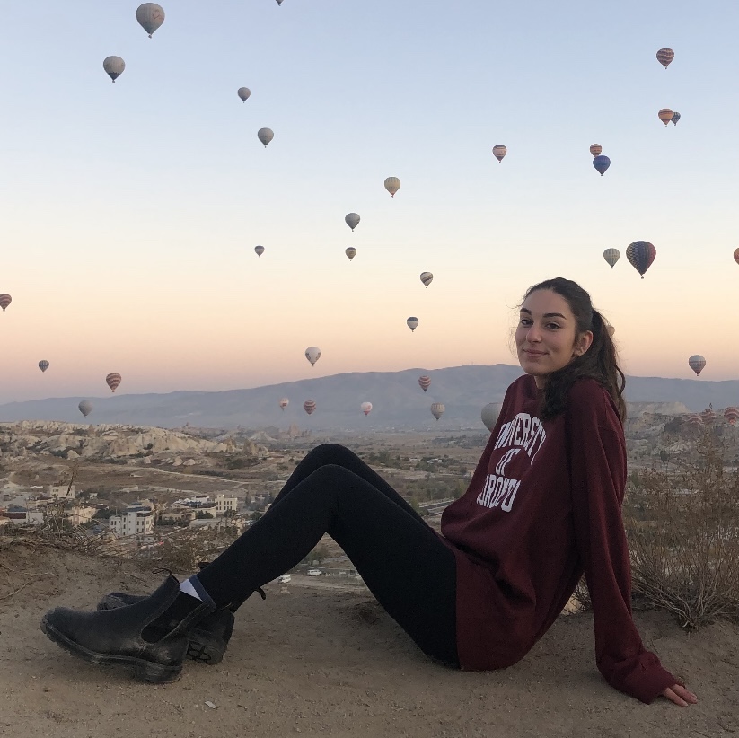 Nicole sitting and smiling on a ledge, while in the background hot air-balloons take off in front of a mountain.