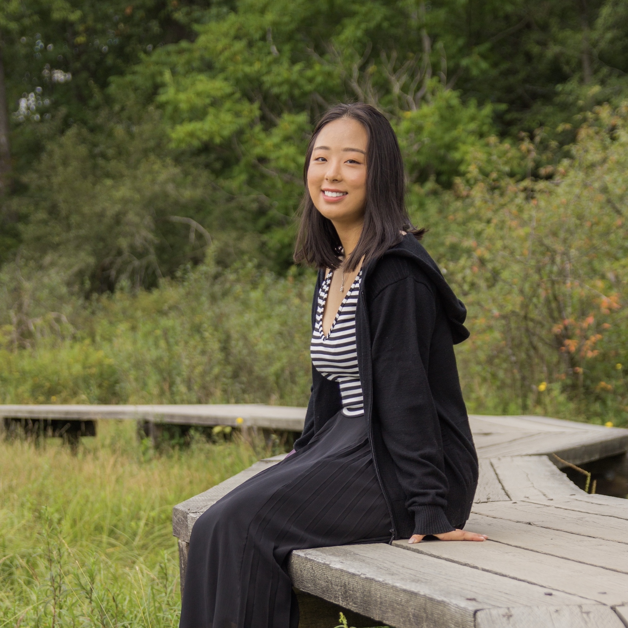 Mingxin smiling and sitting on an elevated wooden path surrounded by trees and foliage.