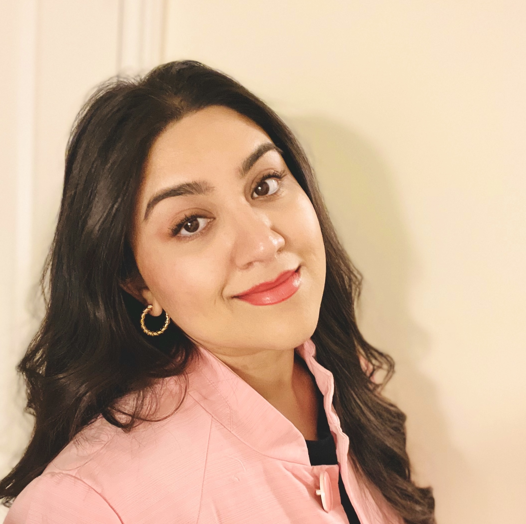 Nida smiling in a pink jacket in front of a white wall. 
