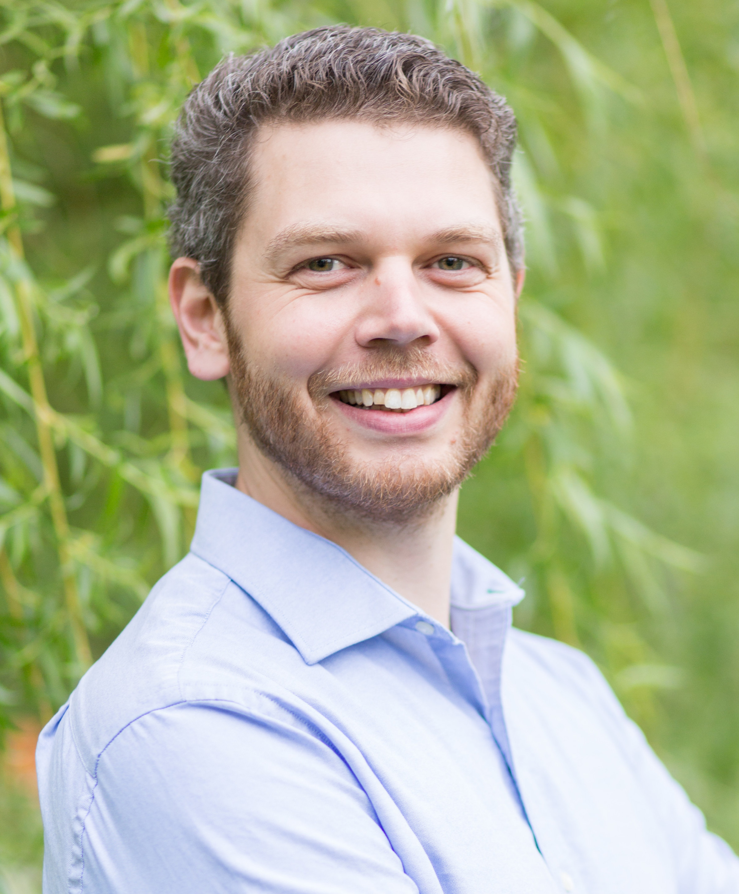 Matthew Adams smiling with arms crossed in front of trees. 