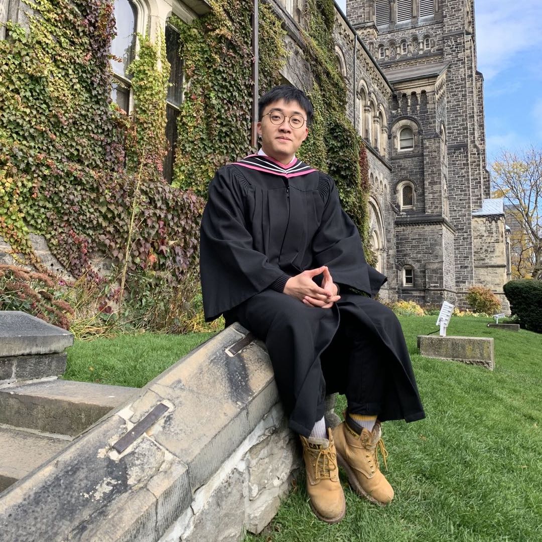 Yihong smiling and sitting in front of the University in his graduation gown.