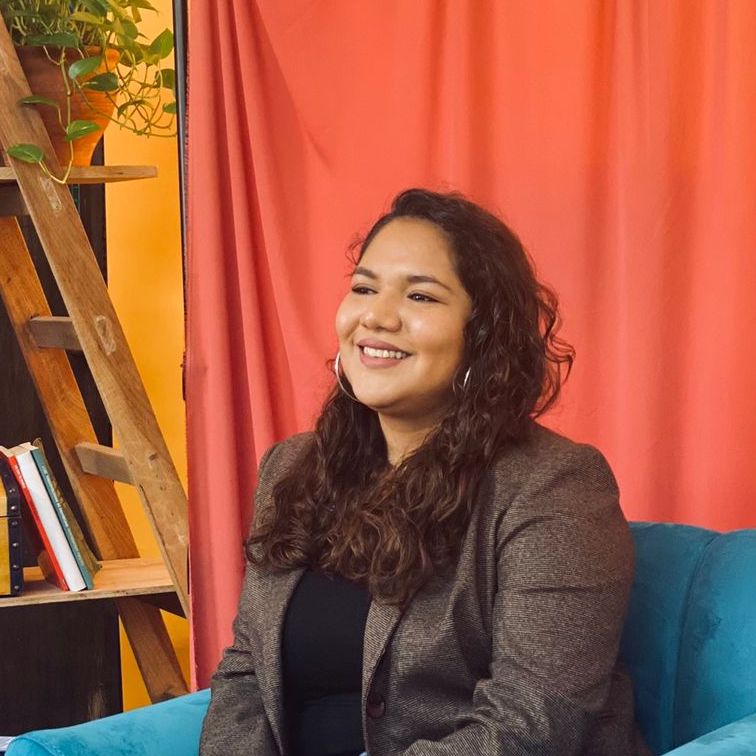 Roxana smiling and sitting in a teal armchair in front of a orange curtain.