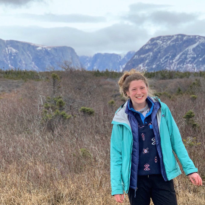 Charlotte smiling and standing in a field in front of a mountain range.