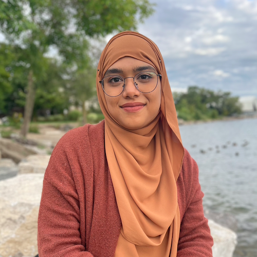 Naziha smiling and sitting on a rock by a beach. 