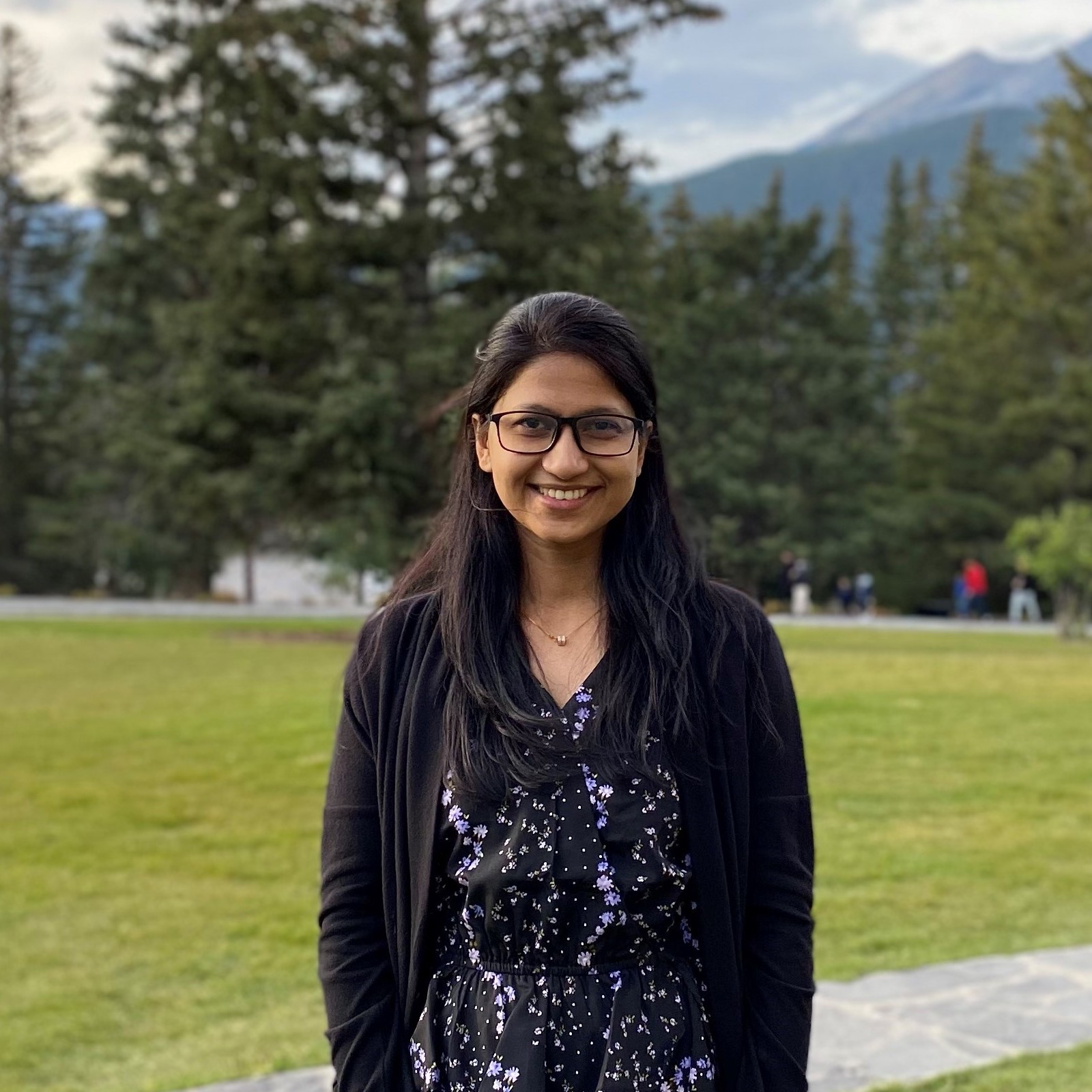Syeda standing and smiling in front of some grass and trees and with mountain in the background. 