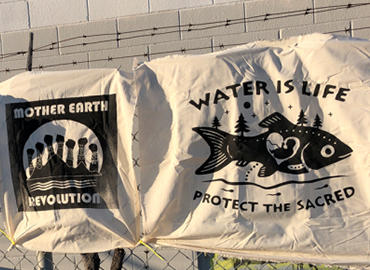Screen printed sheets hung on barbed wire fence. One reads &amp;#039;Water is Life, protect the sacred&amp;#039; the other reads &amp;#039;Mother Earth Revolution&amp;#039;