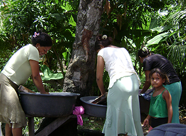 two women bending over wheelbarrows in a forested area. a child stands in front of them, looking at the camera