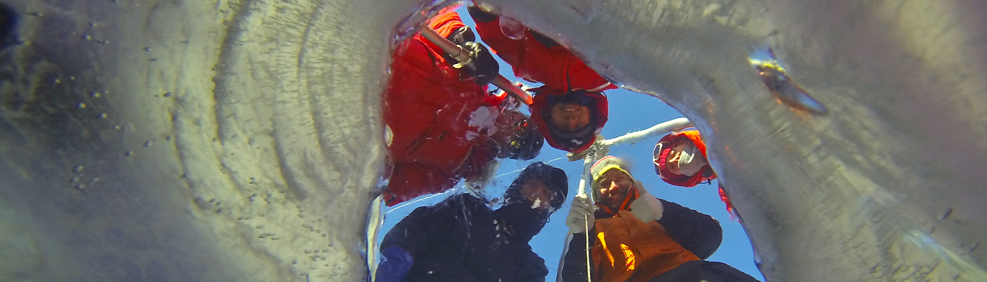 A photo taken by a camera beneath ice of three researchers peering into the ice. 
